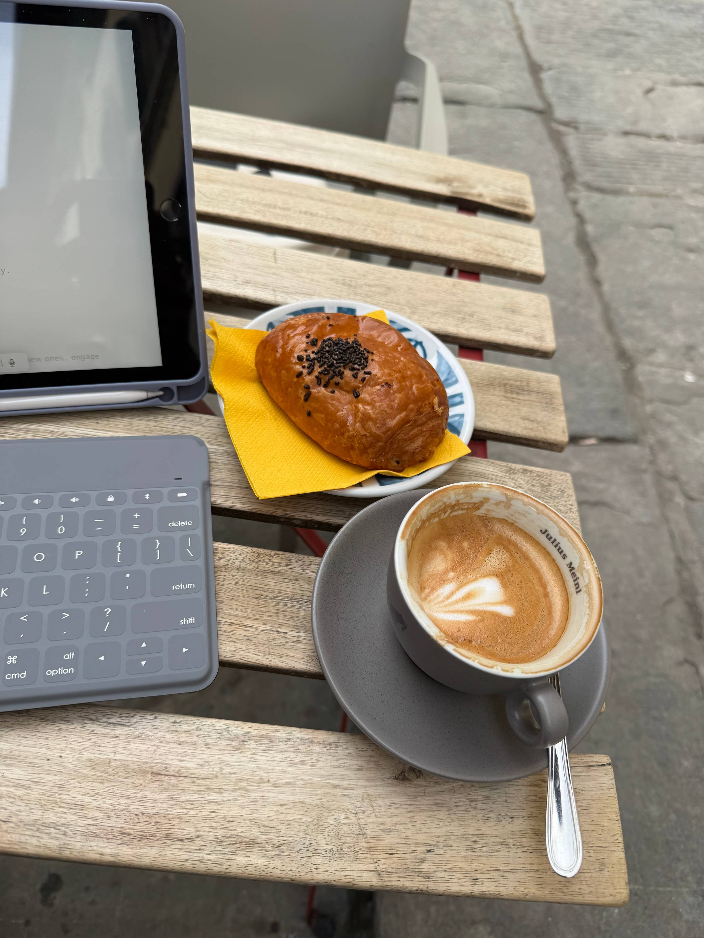 Overhead view of an outdoor café table with a Julius Meinl cappuccino, a chocolate-chip-topped brioche bun on a small plate, and an iPad with Bluetooth keyboard, set on a slatted wooden table against a stone-paved street.