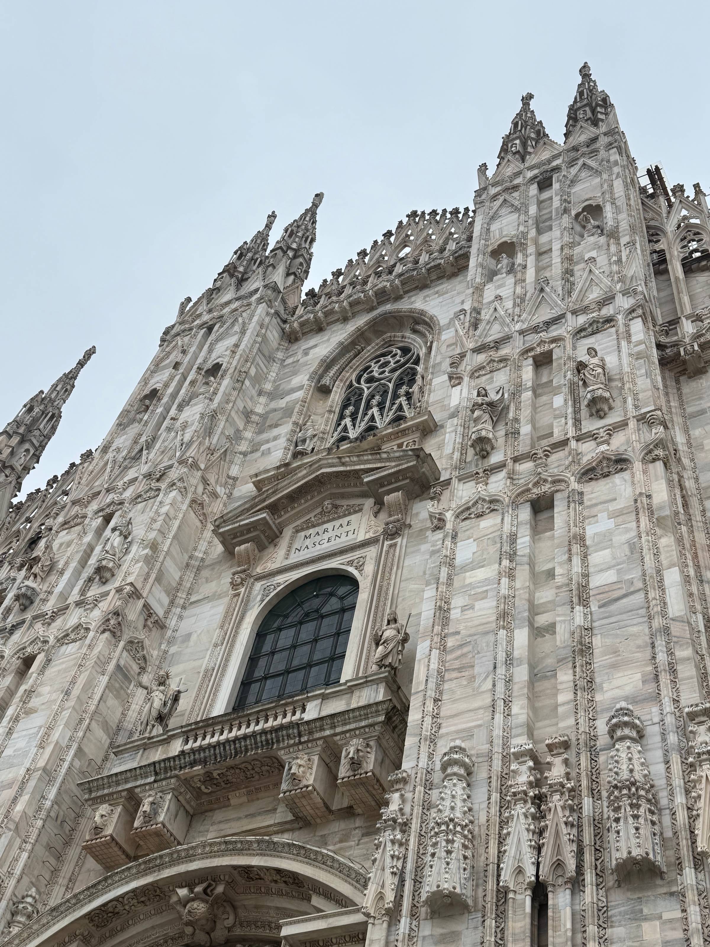 Upward view of the Milan Duomo's white marble Gothic façade, showing "Mariae Nascenti" inscribed above an arched window, surrounded by intricate pinnacles, statues, and decorative stonework against a pale sky.
