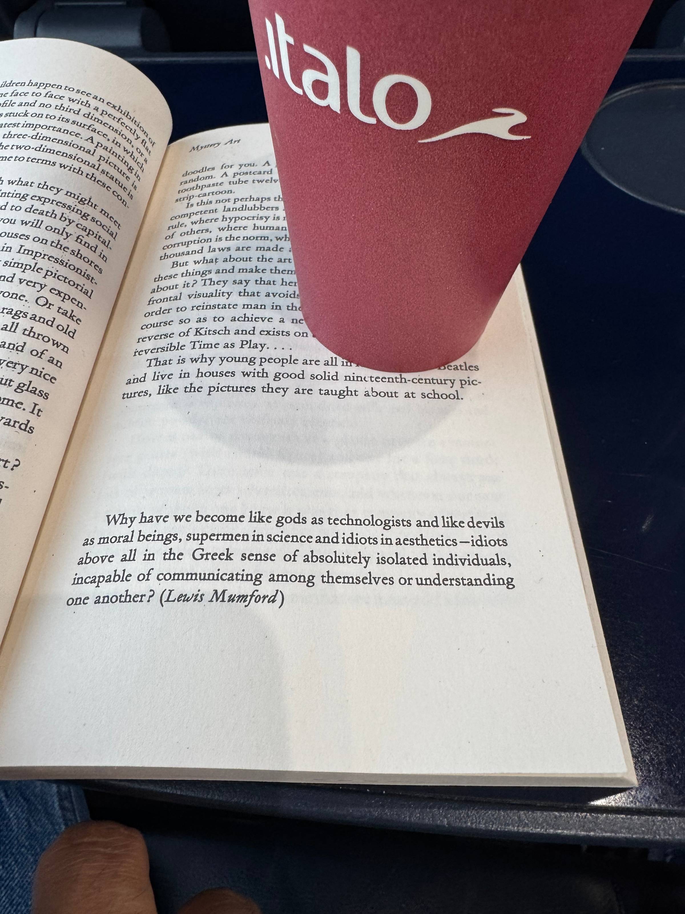 Open book on a train table beside a red Italo train coffee cup, showing a Lewis Mumford quote in italics: "Why have we become like gods as technologists and like devils as moral beings, supermen in science and idiots in aesthetics..."