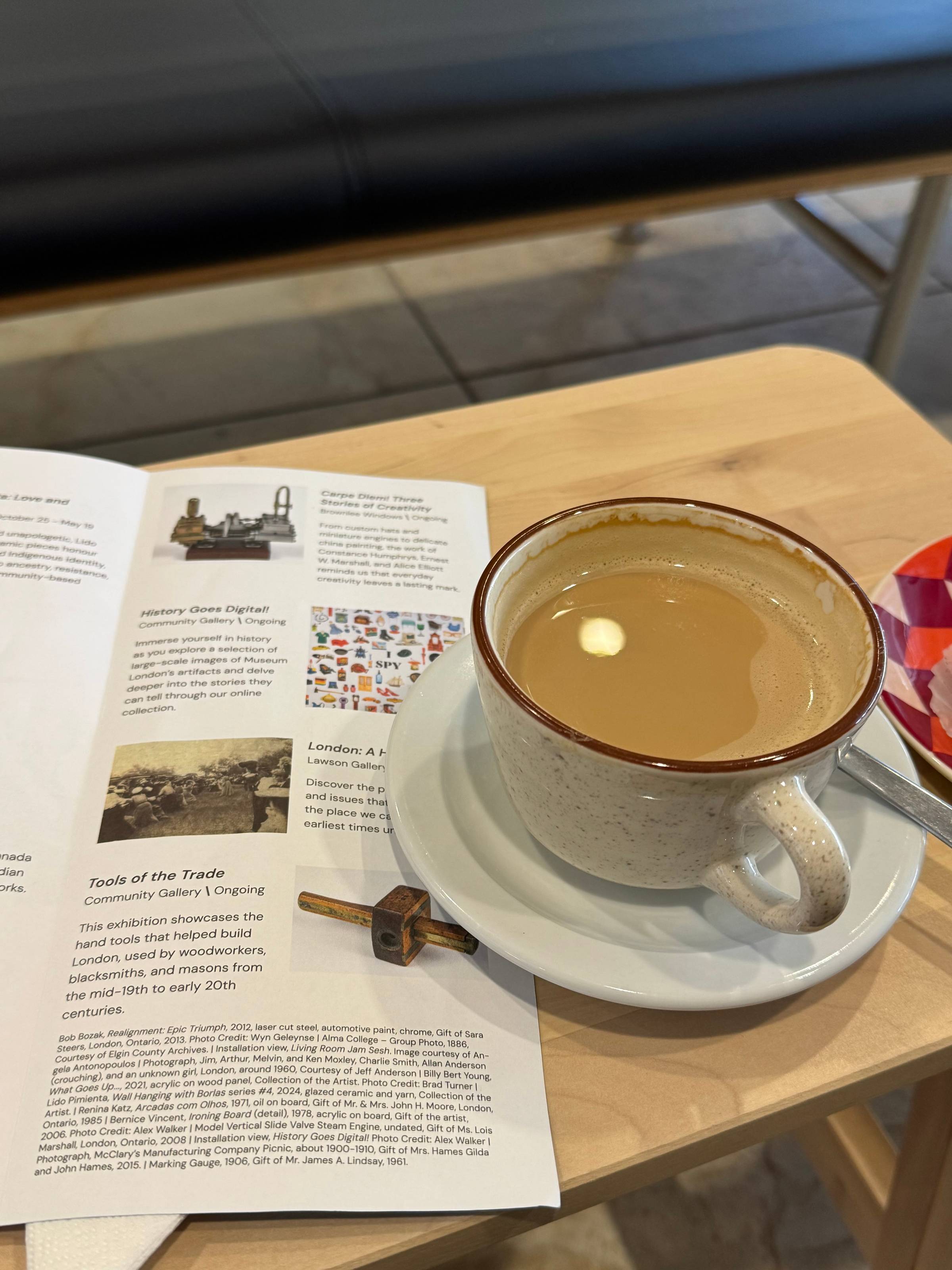 A latte in a brown-rimmed ceramic cup on a saucer, resting on a gallery guide showing current exhibitions including "Tools of the Trade," "History Goes Digital," and "Carpe Diem! Three Stories of Creativity." A small antique marking gauge sits on the brochure beside the cup