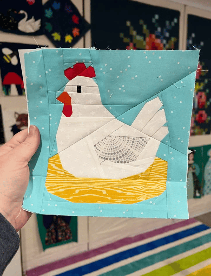a foundation paper pieced block featuring a white chicken in a nest on a blue background held up by the quilter in front of a design wall