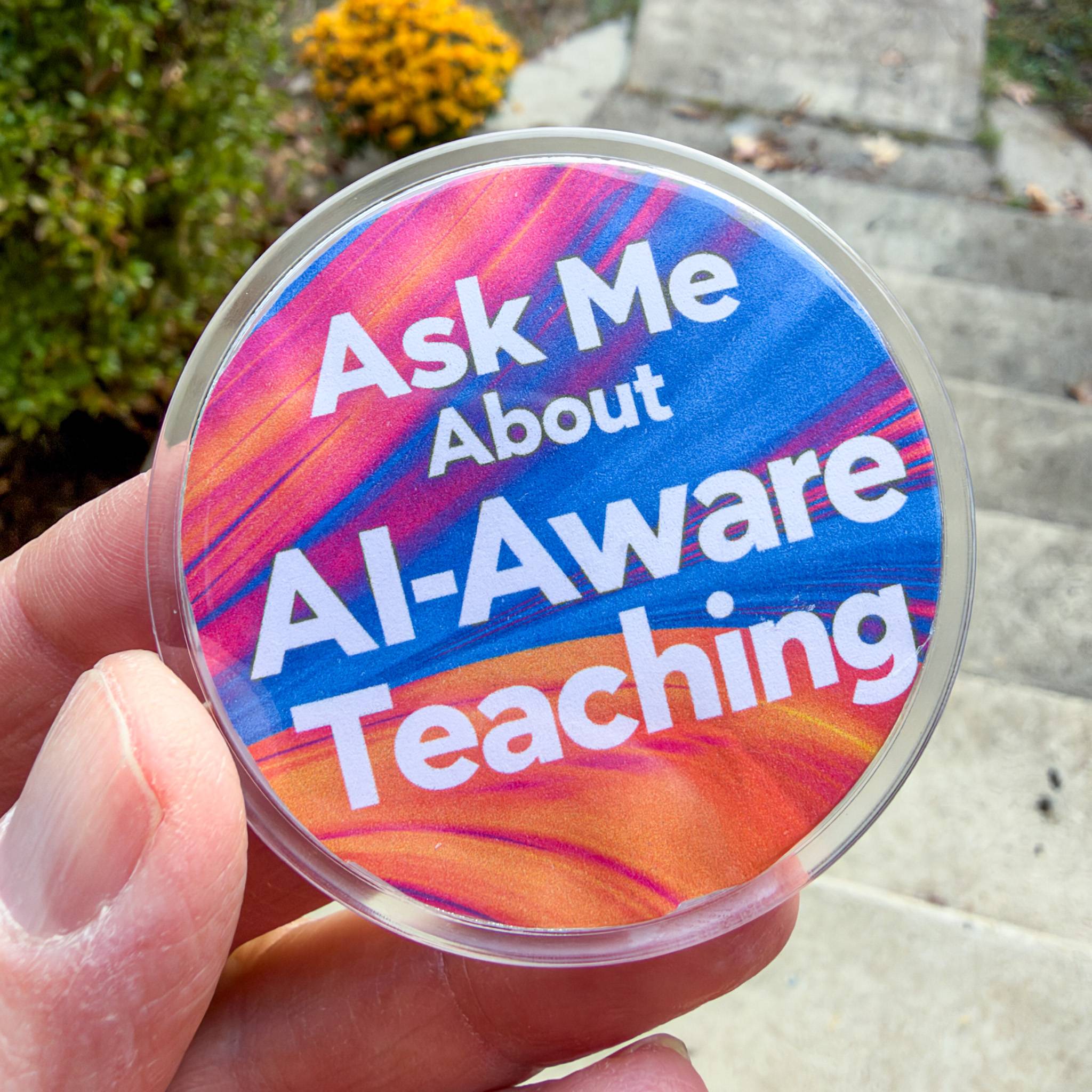 My hand holding a homemade button reading "Ask me about AI-aware teaching" using the font and colors of the book cover