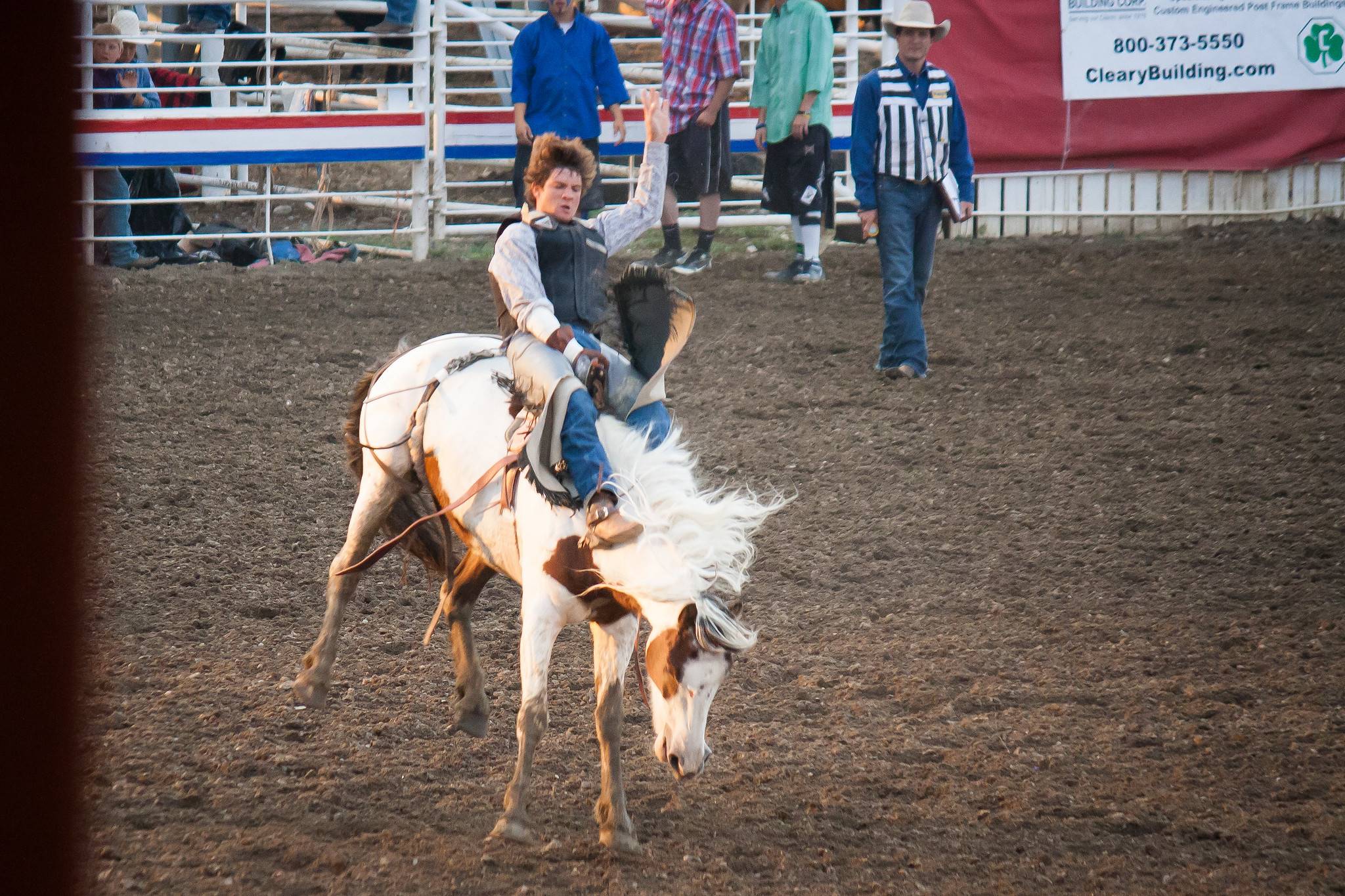 Photo of a young adult in cowboy attire riding a bucking bronco, one hand on the saddle, one in the air, and a look of intense focus on his face.