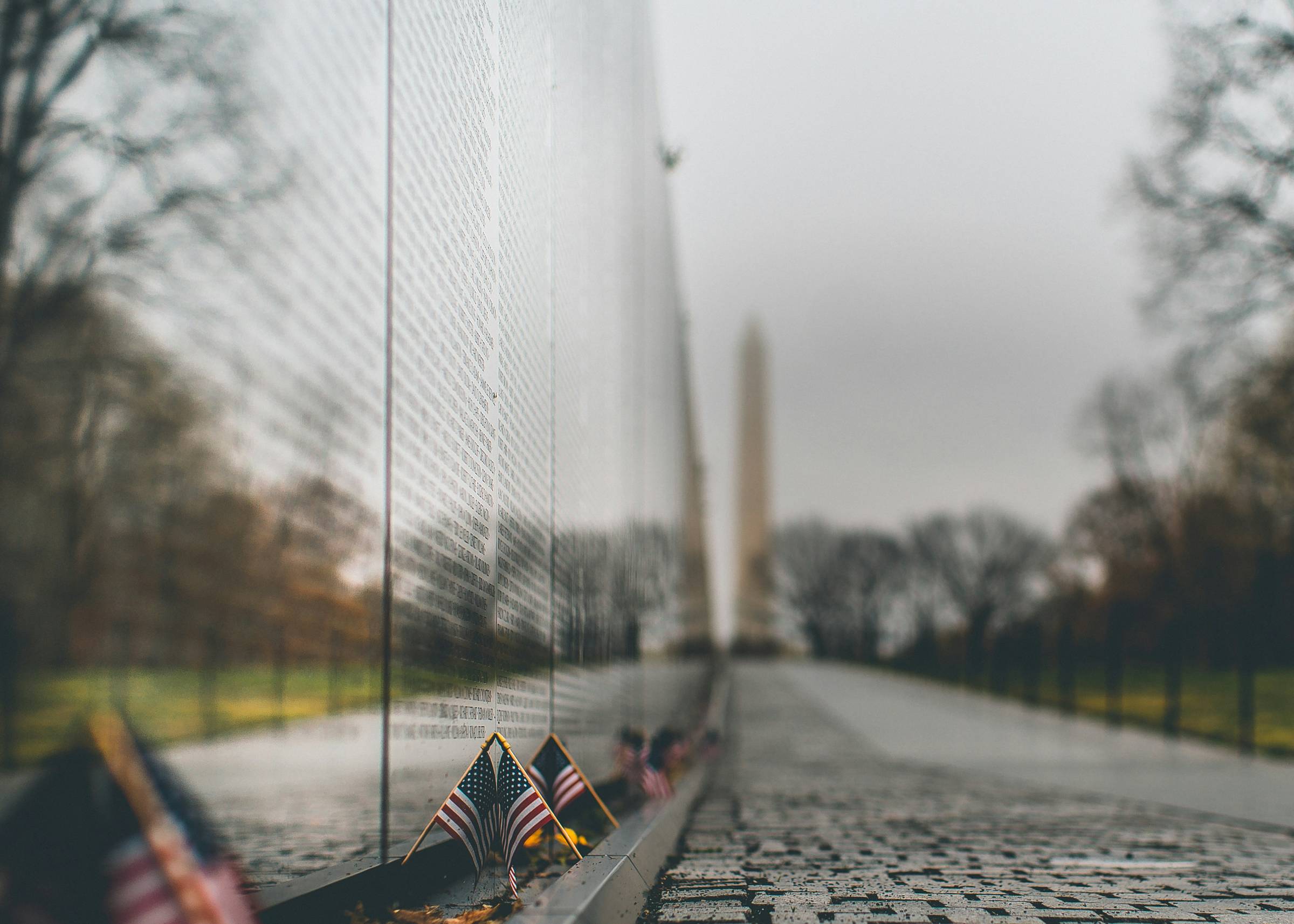 A close up of the Viet Nam wall.  There are small American Flags at the base of the wall, and a blurred image of the Washington Memorial in the background.