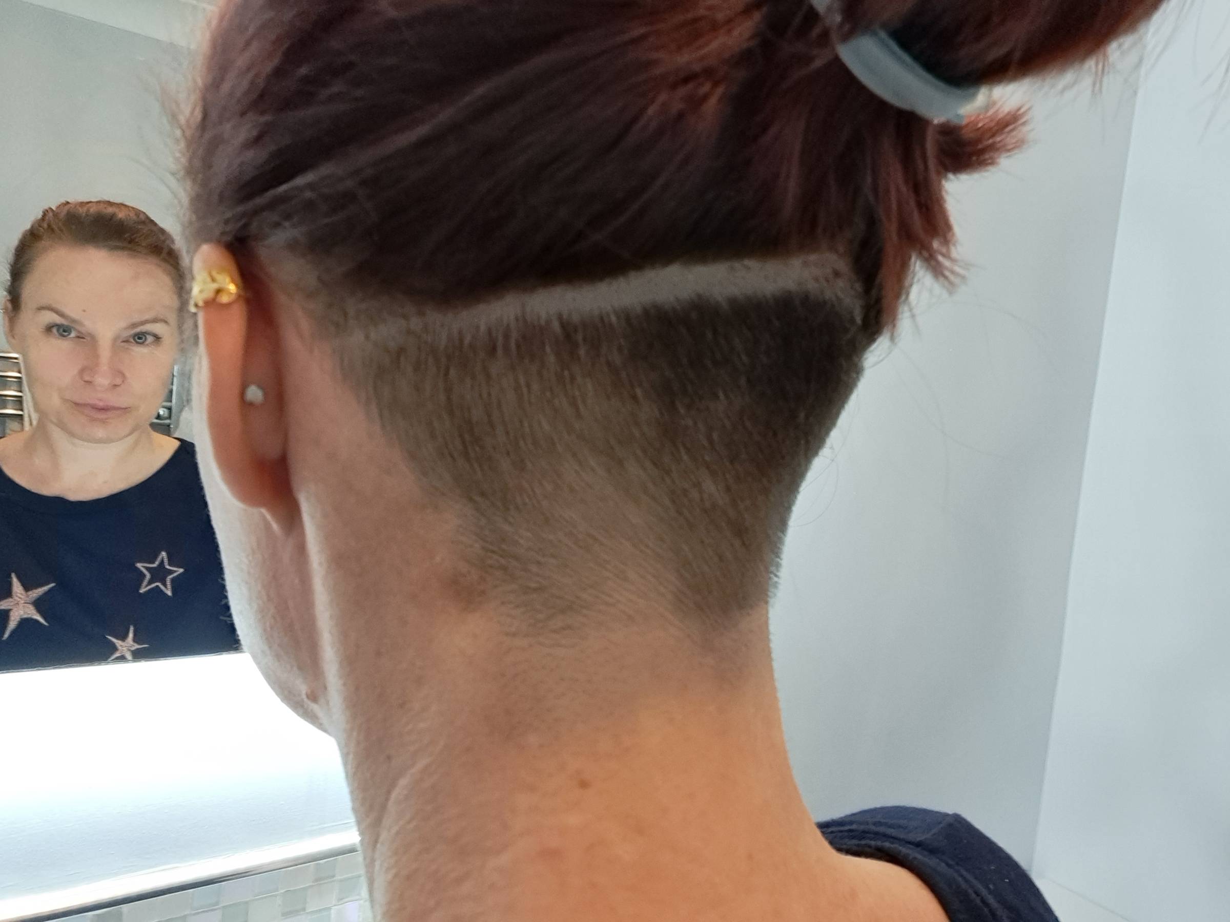 LA person with their hair tied up, showing the back of their head with a freshly shaved undercut. The shaved section extends up from the nape of the neck in a neat V-shape, with a clean horizontal line separating the shaved area from the longer hair above. The person’s reflection is visible in the mirror, showing them wearing a dark top with star patterns.