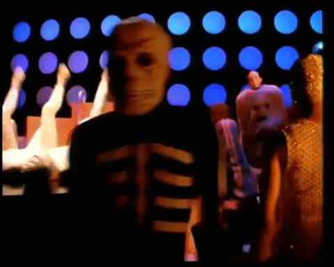 A looping video shows people dancing in costumes against a backdrop of large blue dots on a black wall. In the foreground, a person in a skeleton suit and skull mask walks toward the camera. To the left, several dancers kick their legs in the air while lying on a raised platform. Other figures in colorful, patterned outfits move in the background, giving the scene a surreal, playful, and slightly eerie vibe.
