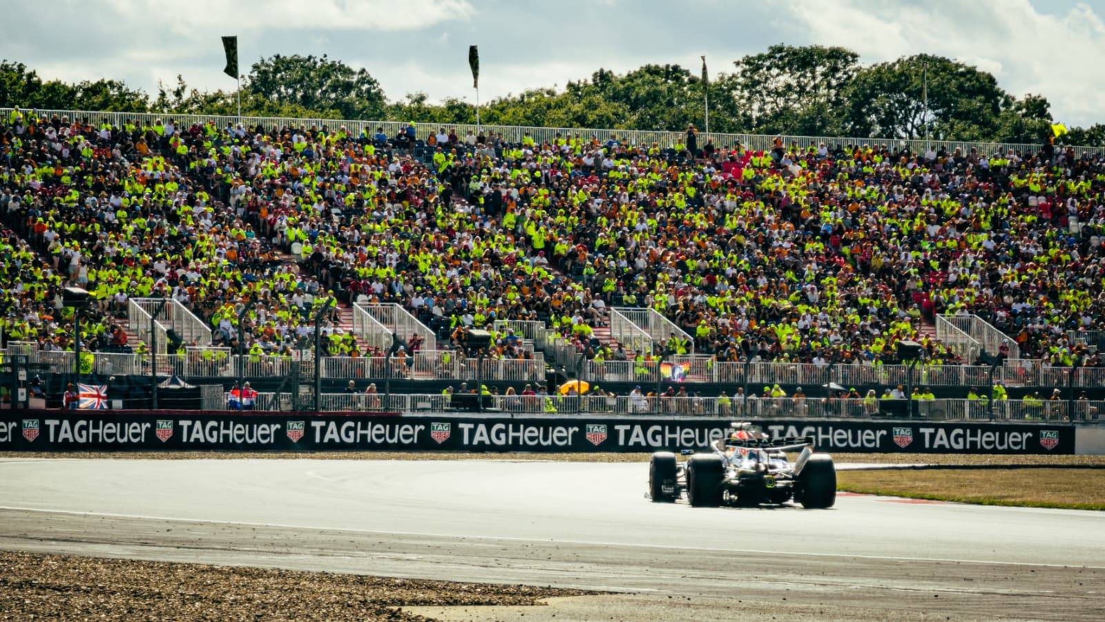 A Formula 1 race car speeds down a track past a massive, packed grandstand under a partly cloudy sky. The crowd is a dense sea of spectators, many wearing bright yellow or neon shirts, and the track barrier features prominent, repeating TAG Heuer branding.
