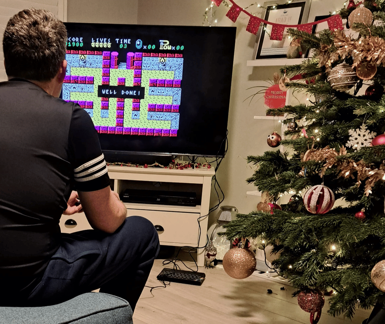 A person sits on a sofa playing a retro video game on a TV, with a ‘Well done!’ message on screen; a decorated Christmas tree stands beside the TV in a cosy living room.