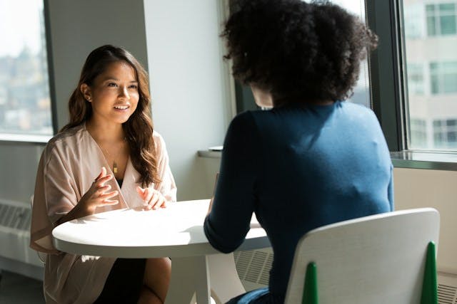 Two women sit at a table facing each other. One is a young Asian woman talking to another woman who we can only see from the back. Her black curly hair hints that she's African-American. The picture suggests a coaching relationship.