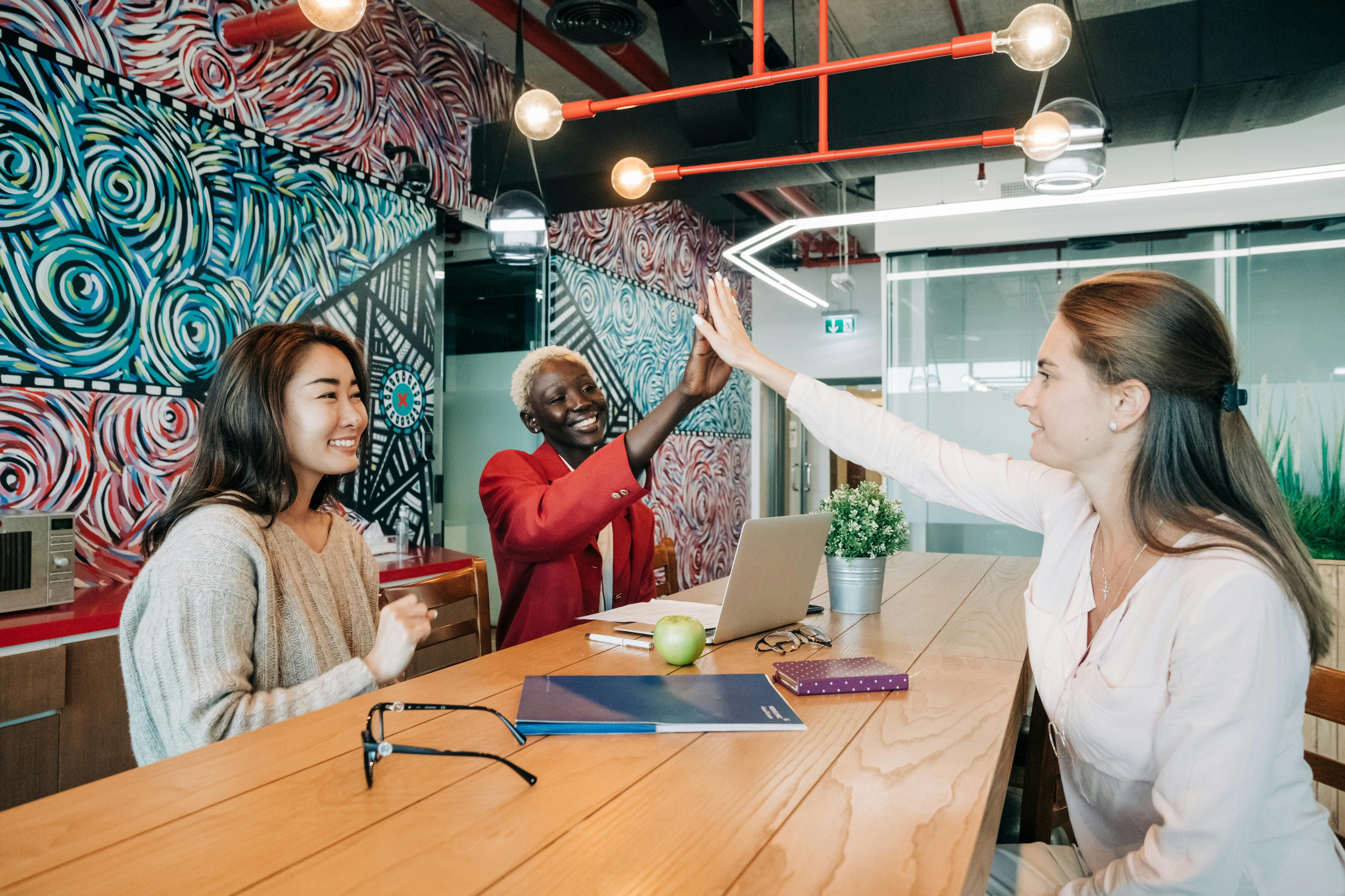 Three women on their 20-30s of different ethnicities sat around a table smiling. Two of them are high fiving each other.