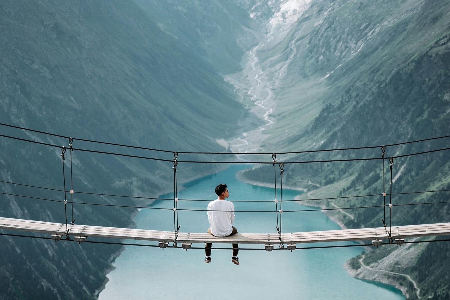 Photo of a man sitting on bridge, looking at mountains.