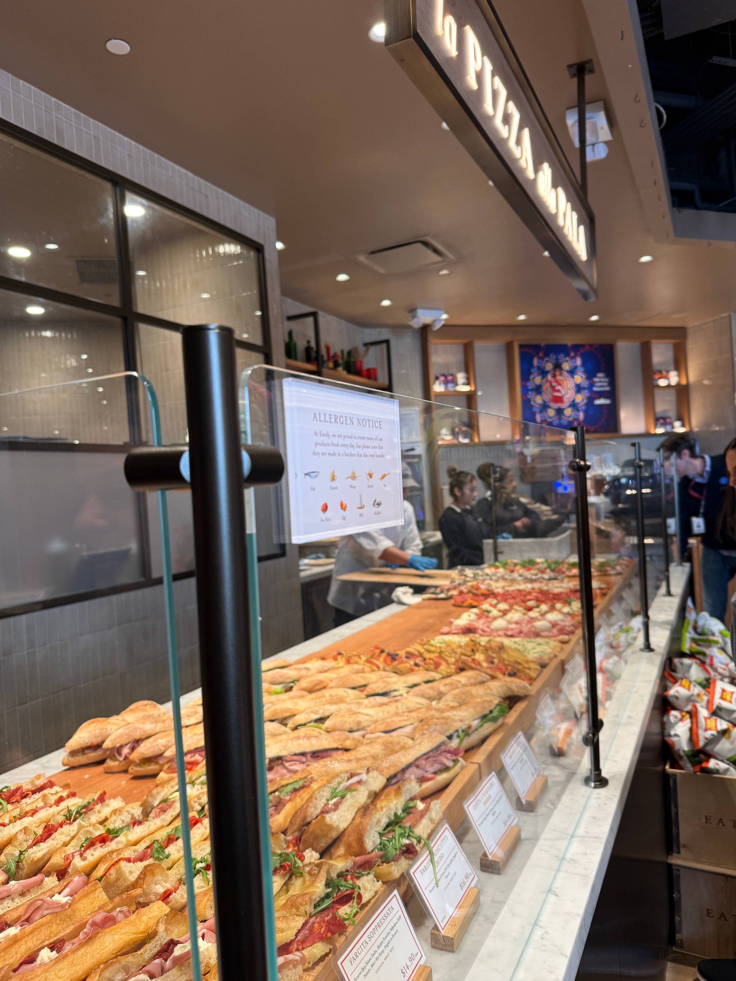 Italian sandwiches displayed behind a glass counter at Eataly, with assorted meats, cheeses, and fresh bread prepared by staff in the background.
