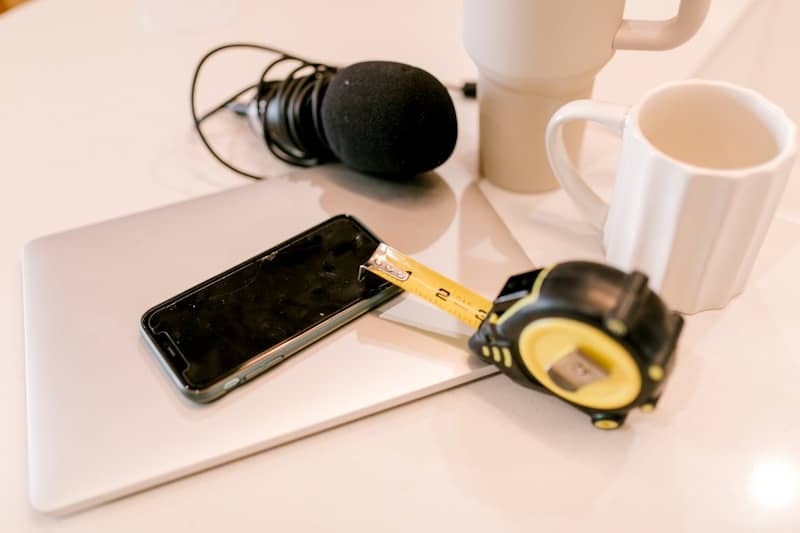 Smartphone, laptop, tape measure, microphone, and mugs on a desk.