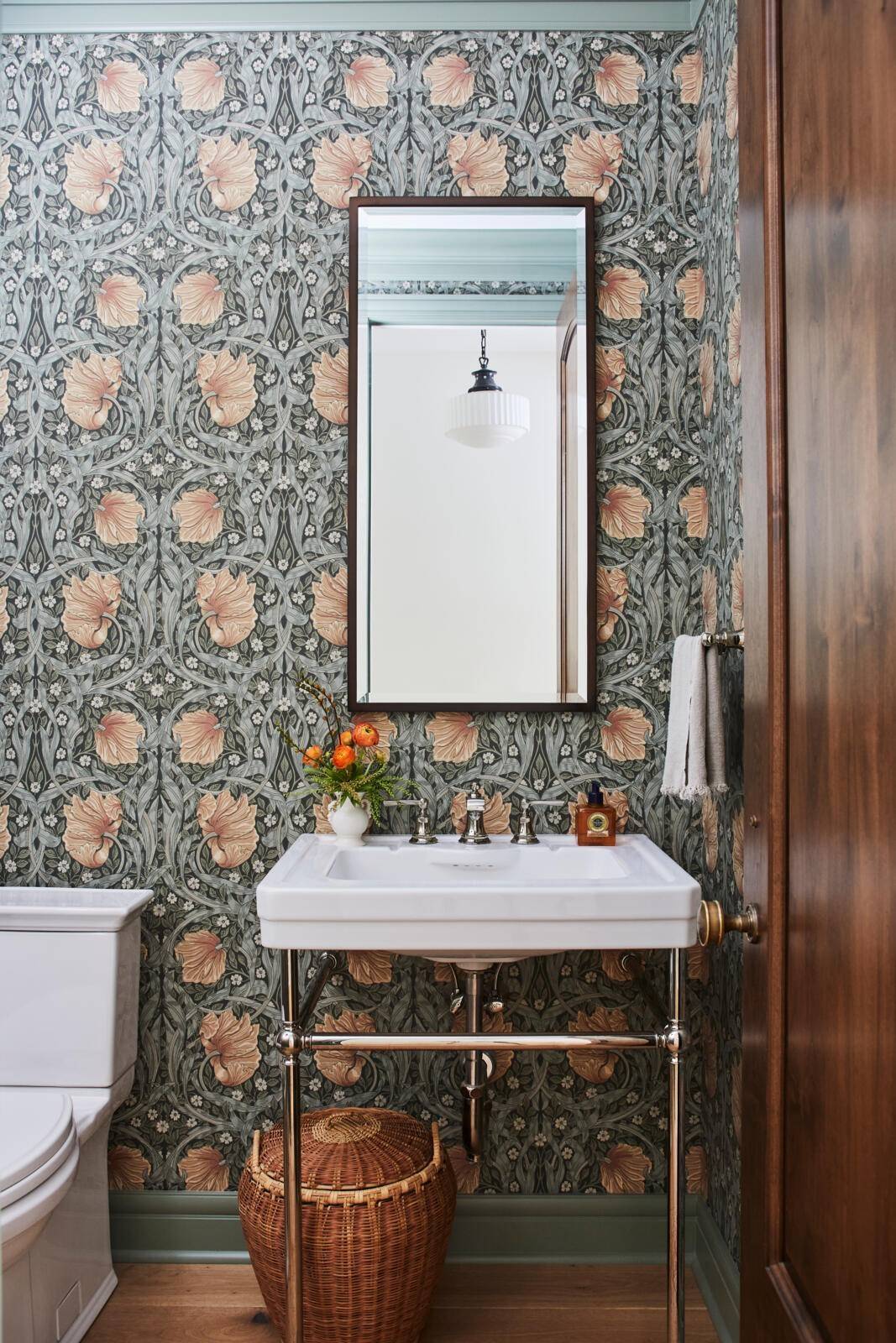 A small bathroom with floral wallpaper in shades of green, pink, and gray. The design features a white pedestal sink with silver fixtures and a rectangular mirror above it. Next to the sink, there's a white towel hanging on a rack. A wicker basket is placed under the sink. To the left of the sink is a white toilet. A vase with orange flowers sits on the sink countertop. The room has wooden floors and a wooden door partially visible on the right.