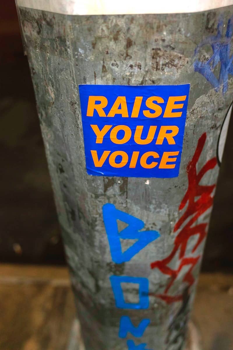 Blue sticker with orange text "raise your voice" on pole