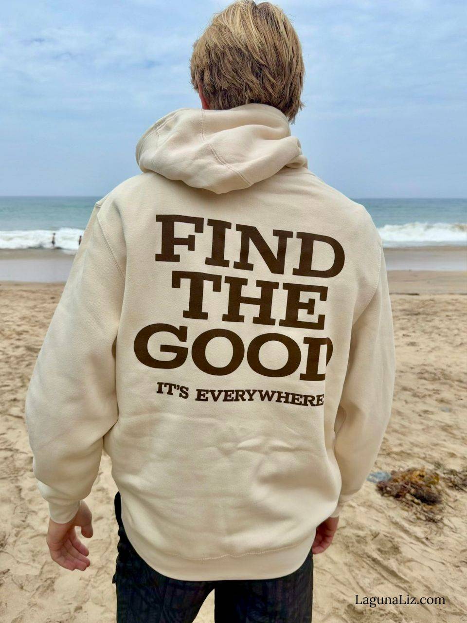 Young man wearing a hooded sweatshirt with the words "Find The Good: It's Everywhere" printed on the back. He is facing the ocean on a sandy beach.