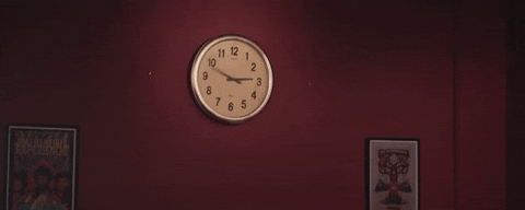 A white, round analogue clock sits on a dark red wall. Its hands spin rapidly as it slowly starts to melt.