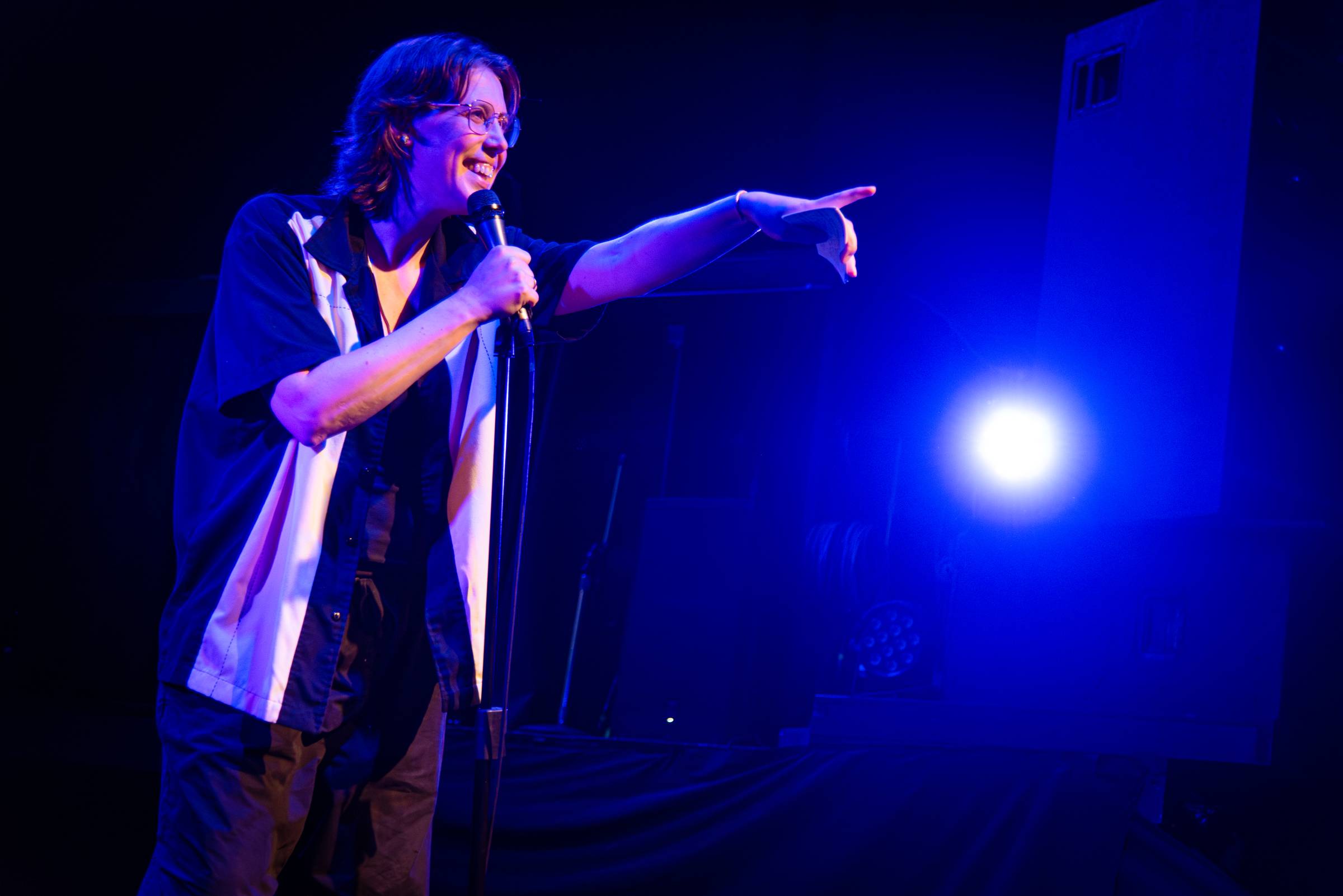 Caucasian androgenous person with brown mullet and glasses points to an audience outside the photo, while holding a microphone.