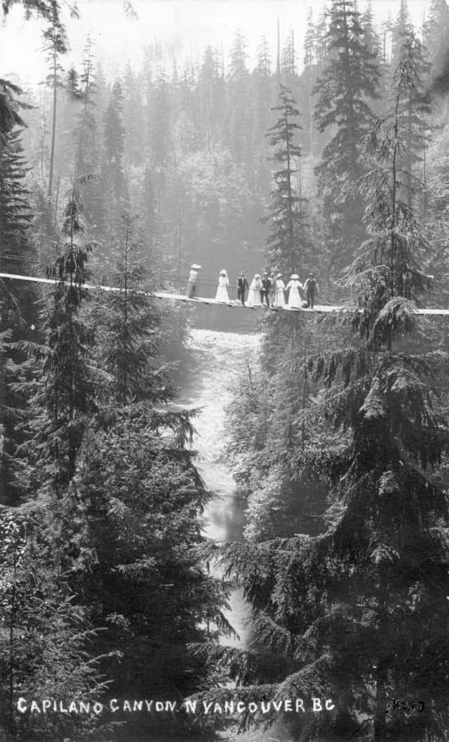 A historical photo of 8 people standing on a long suspension bridge over tall fir trees and a river canyon at Capilano.