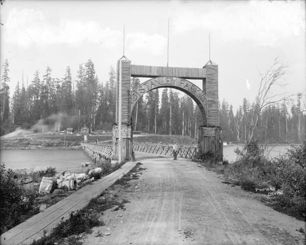 Photo of Stanley Park entrance in 1890s. Features a wooden arch with park trees in background.
