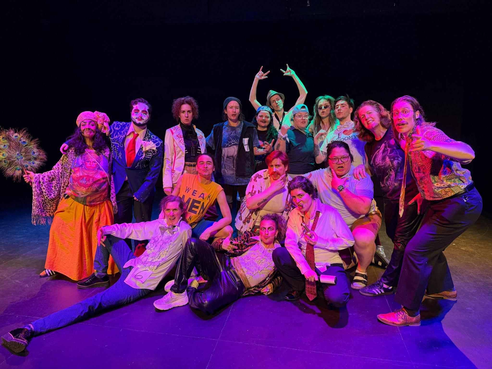 A group of 17 drag performers pose for a photo, smiling with fake facial hair,under red stage lights.