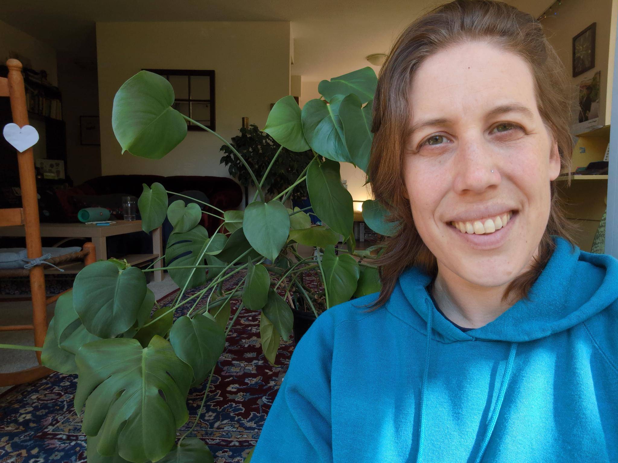 A caucasian 40 year old femme person smiles at the camera in front of a monstera plant.