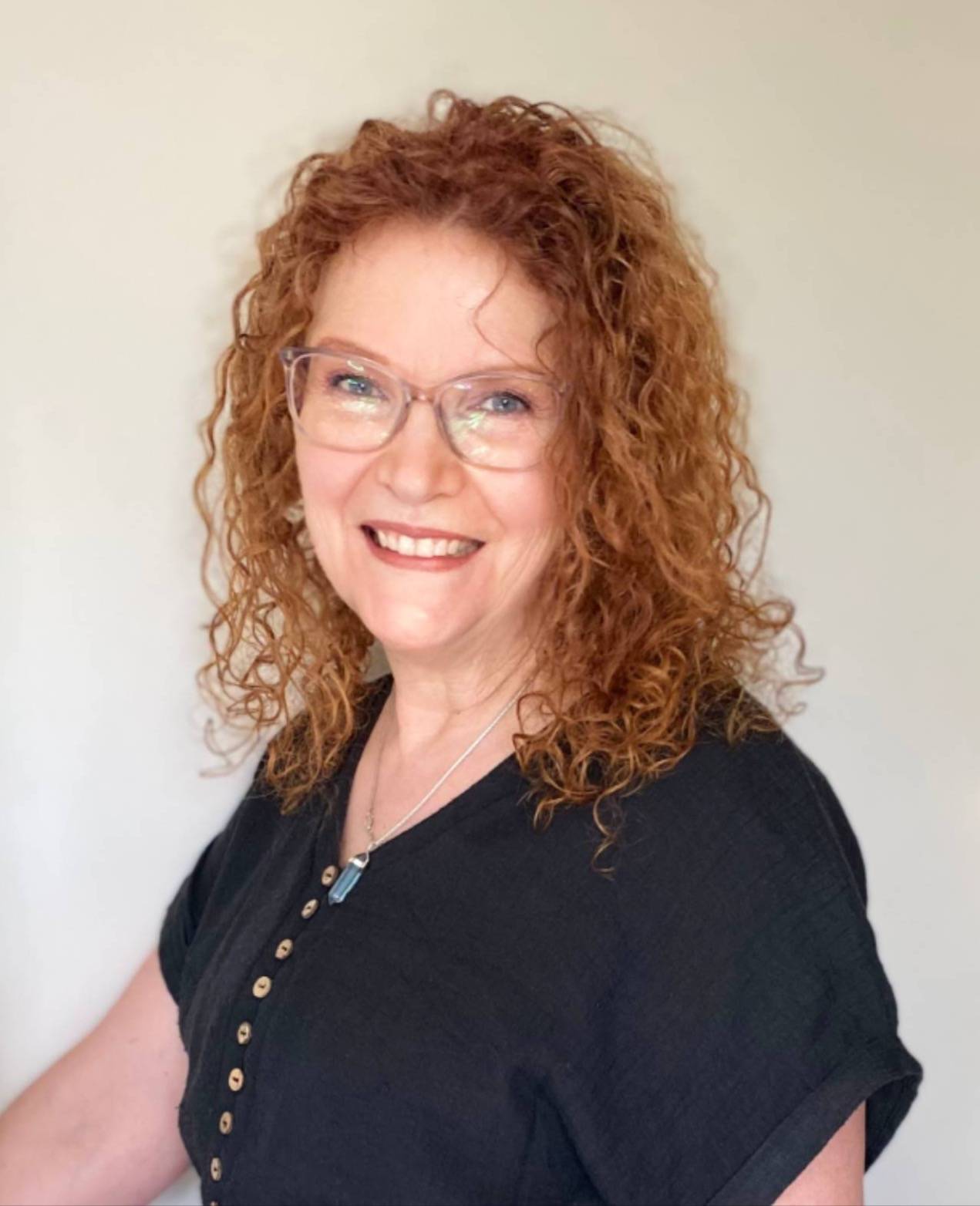 photo of a smiling woman with glasses and curly auburn shoulder length hair, wearing a dark navy short sleeved top