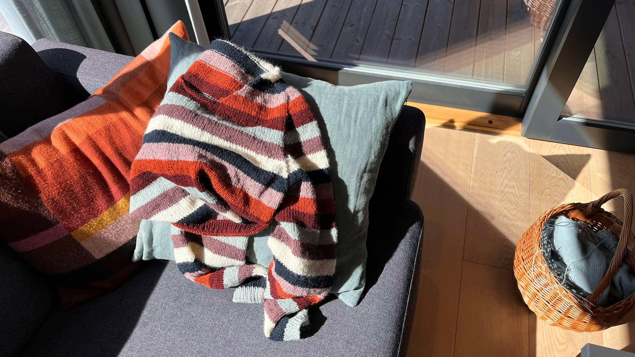 A brightly striped sweater is lying crumpled on a sofa cushion, bright sunlight shines on it.