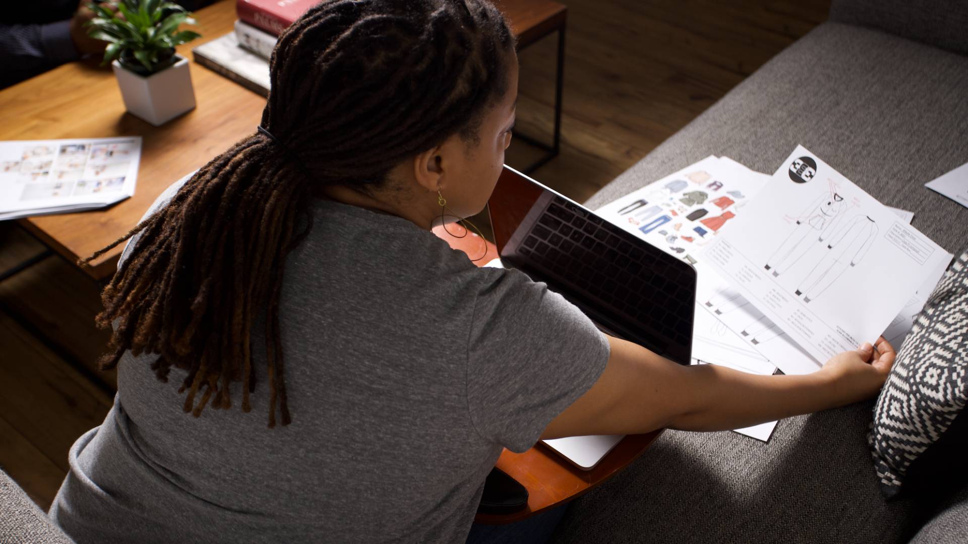 A person with long locs pulled back works at a laptop on a couch, reviewing a printed tech pack with a fashion flat sketch and spec details. A trim and materials sheet sits nearby on the coffee table alongside a small potted plant and stacked books.