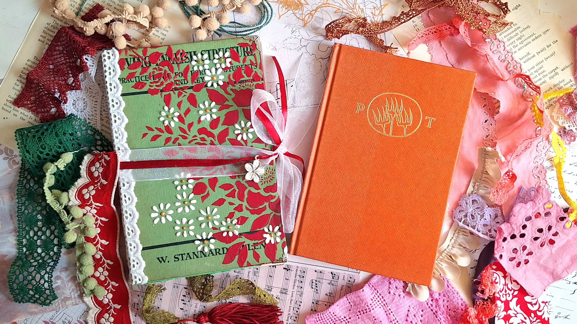 Overhead view of a vintage-style journal and an old book surrounded by colorful trims, lace, ribbons, and fabric scraps on a creative workspace. The journal has a green cover decorated with red floral stencil and white 3D flowers, tied with sheer and red 