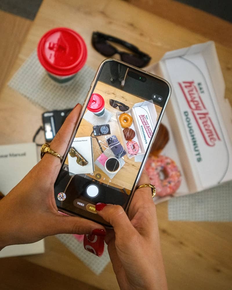 Smartphone capturing an overhead shot of donuts, coffee, and sunglasses.