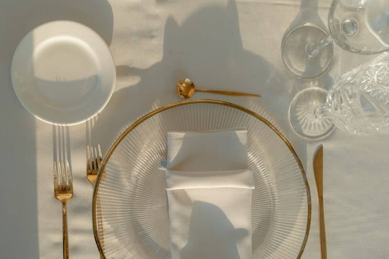 A place setting with a glass plate, gold cutlery, and a white napkin on a tablecloth