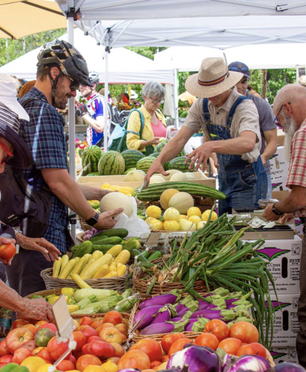 Shoppers and a farmer in overalls and a straw hat browse and sell a colorful array of fresh vegetables and melons at an outdoor farmers market beneath white canopy tents.