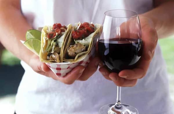 A person in a white shirt holds two fish tacos in a checkered paper holder and a glass of red wine.