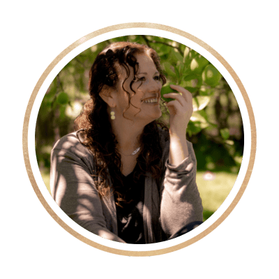 A woman with curly hair is sitting outdoors, surrounded by greenery. She is smiling and looking to her left, her hand raised near her face. She is wearing a dark top and a light sweater. The image has a circular frame with a gold border.