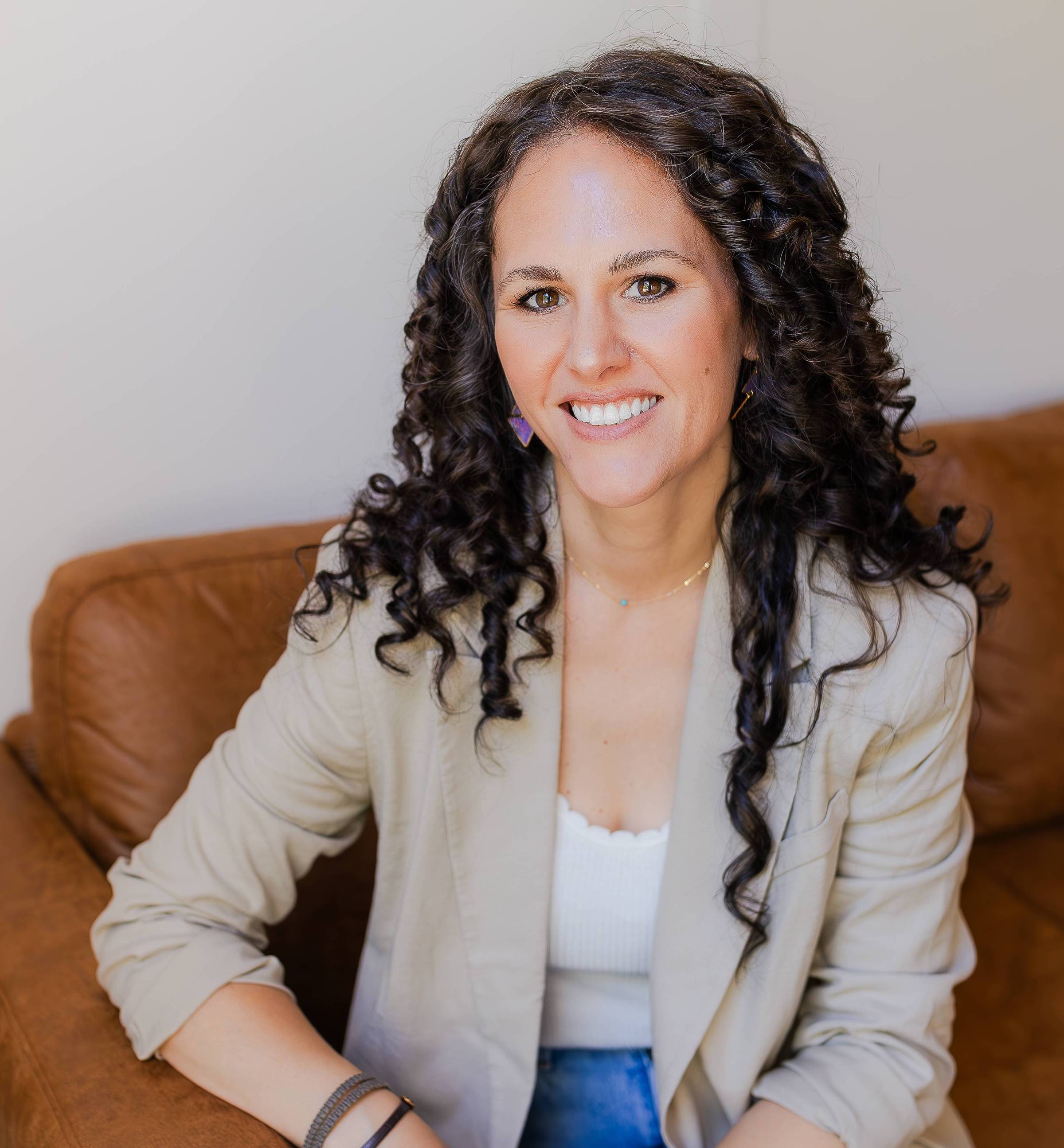A woman with long curly hair is smiling while sitting on a brown sofa. She is wearing a light-colored blazer over a white top, and blue jeans. Her arms are relaxed, one resting on the sofa. She has earrings and a delicate necklace. The background is plain, enhancing her cheerful expression.