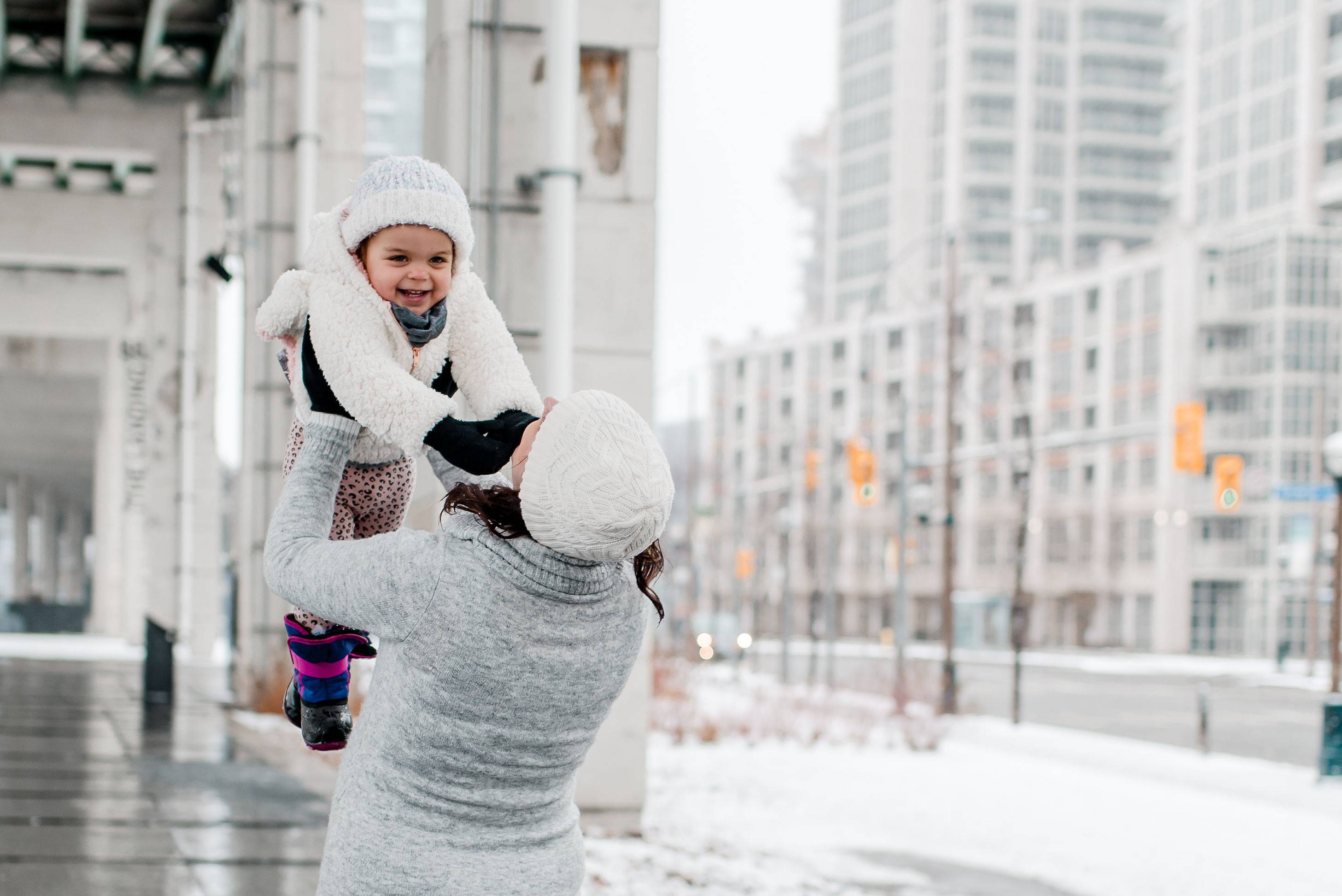  A mother and her baby dressed warmly in winter coats and scarves, walking through a snowy city street with softly falling snow and decorated lampposts in the background. The baby is snug in a carrier, and the mother is smiling warmly.  