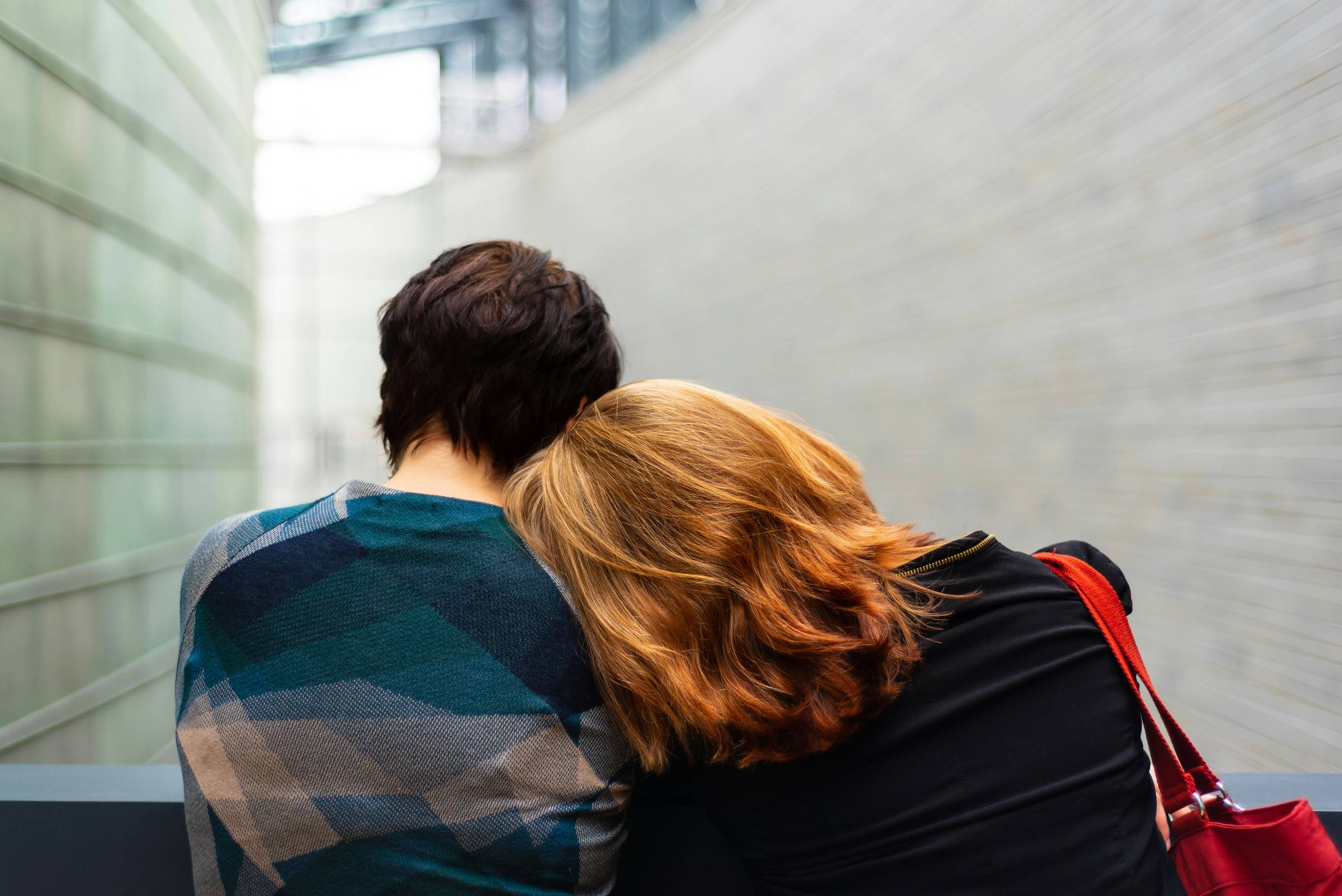 Two women standing side by side, facing away from the camera. One is taller with short brown hair. The other has medium-length red hair, and she is leaning her head on her friend's shoulder.