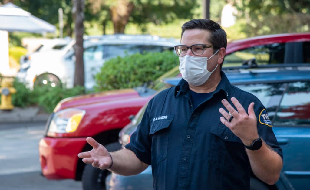 Lt. Gregg, one of the paramedics who responded to Will's sudden cardiac arrest in 2020. In this photo he is wearing a navy blue button down uniform shirt and a surgical mask. He is white, has short brown hair, and wears glasses as well as a smart watch, wristband, and wedding ring.