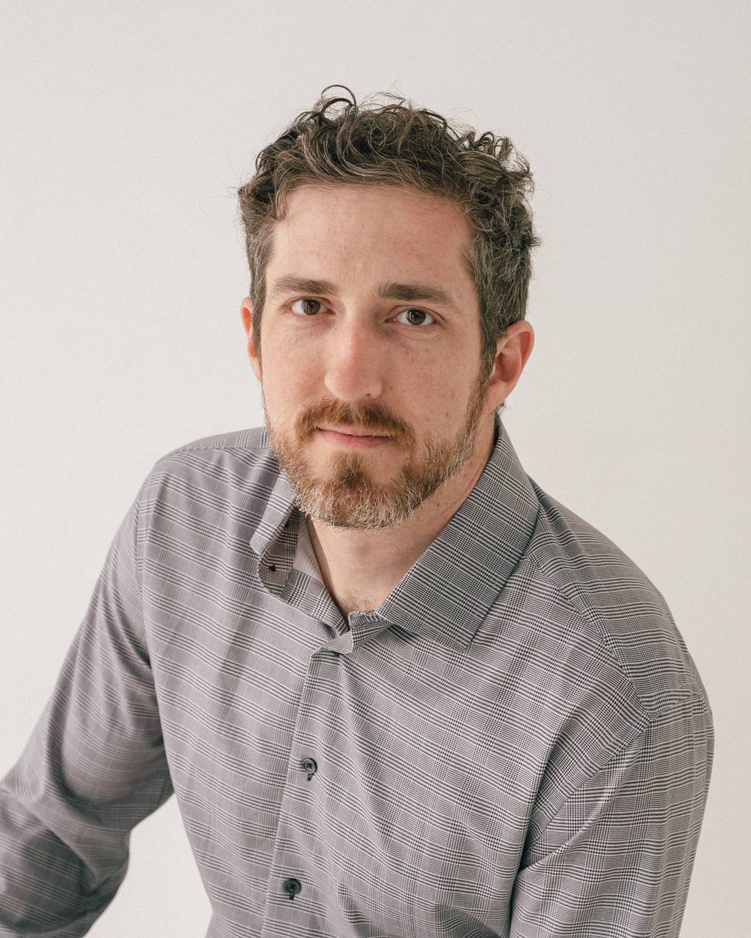 A photo of Will. He has pale skin, brown eyes, short brown & grey curly hair, and a short beard. He's wearing a light grey button down shirt, and he's looking at the camera with a slight, knowing smile.