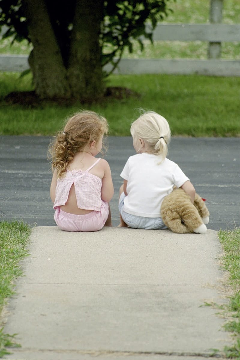 Two little girls sitting on the sidewalk with a teddy bear