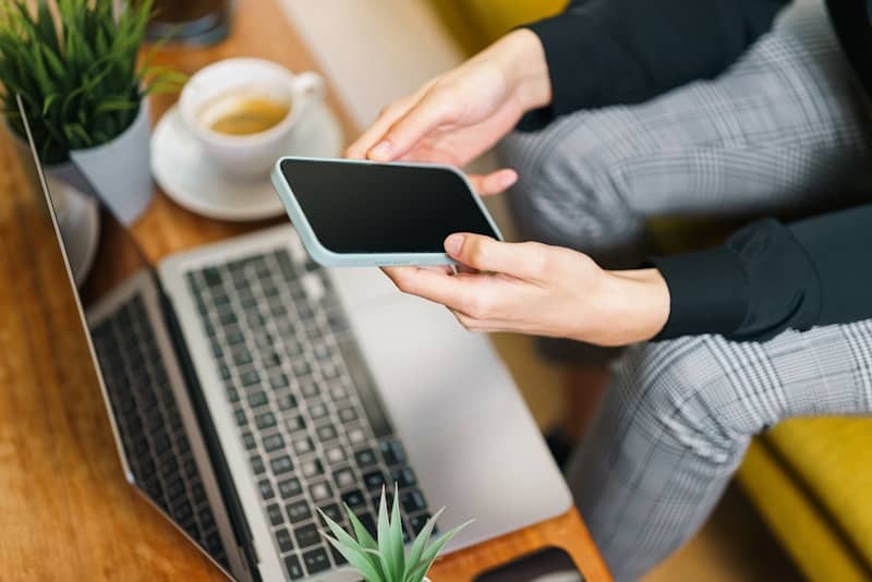 A woman sitting in front of a laptop computer holding a cell phone