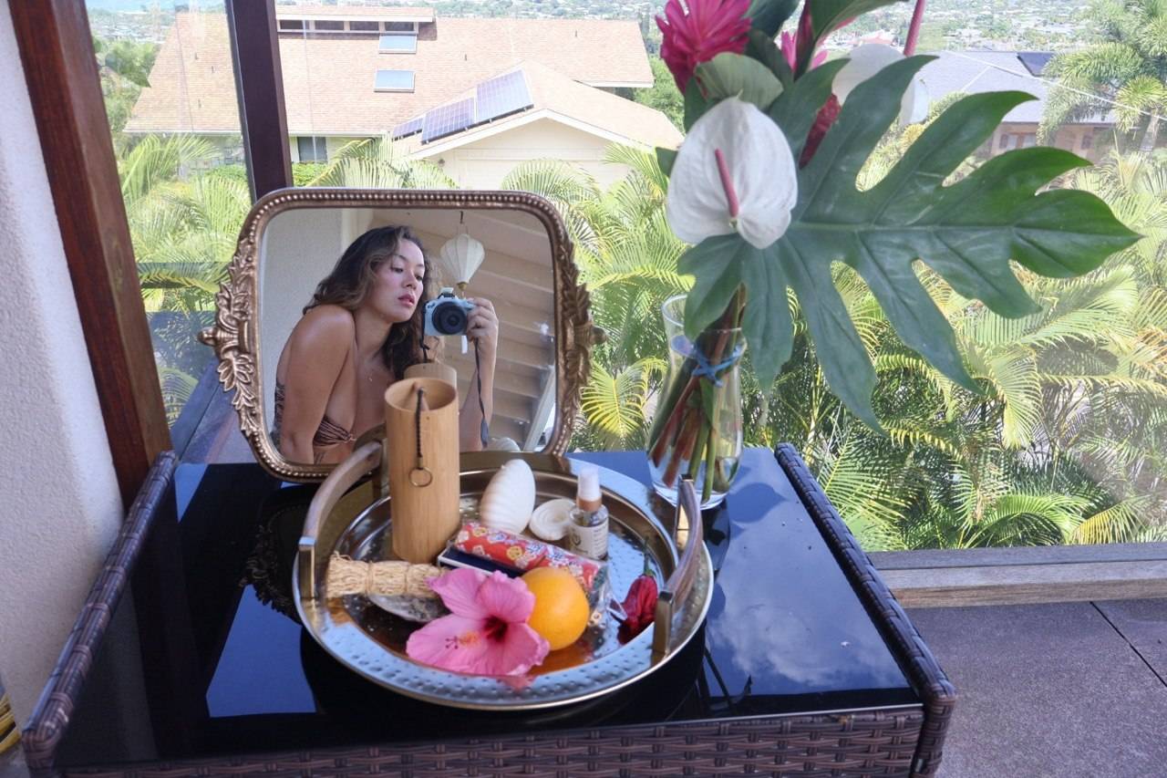 aiko taking a mirror photo overlooking a small altar with plants, anthurium, hibiscus, shells, vetiver bundle, and chimes