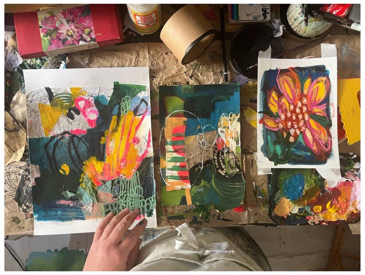 Struggling to start your art? Learn the power of 'dining in' with your creativity. Try our Permission to Play community for free. Image of a desk with three work in progress artworks on a studio table photographed from above with artist, Delphie Joy, rest