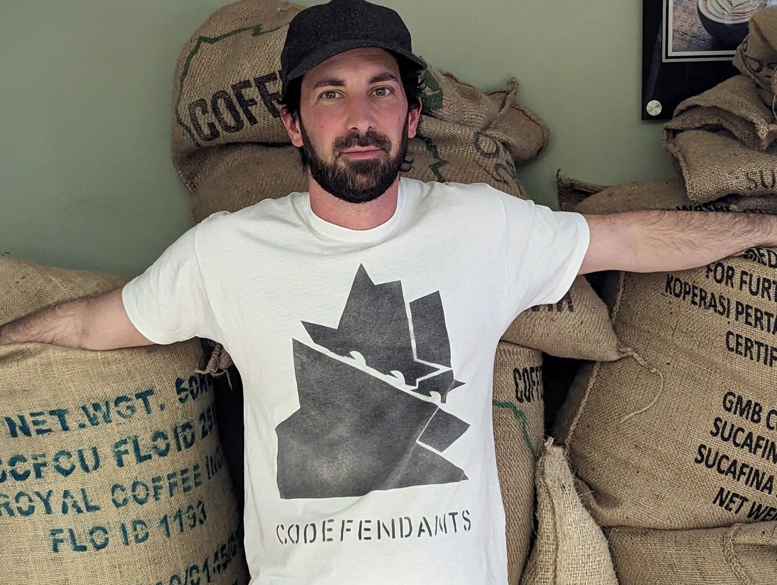 Pepe Bandit with arms open in front of burlap sacks of green coffee, wearing a Codefendants shirt