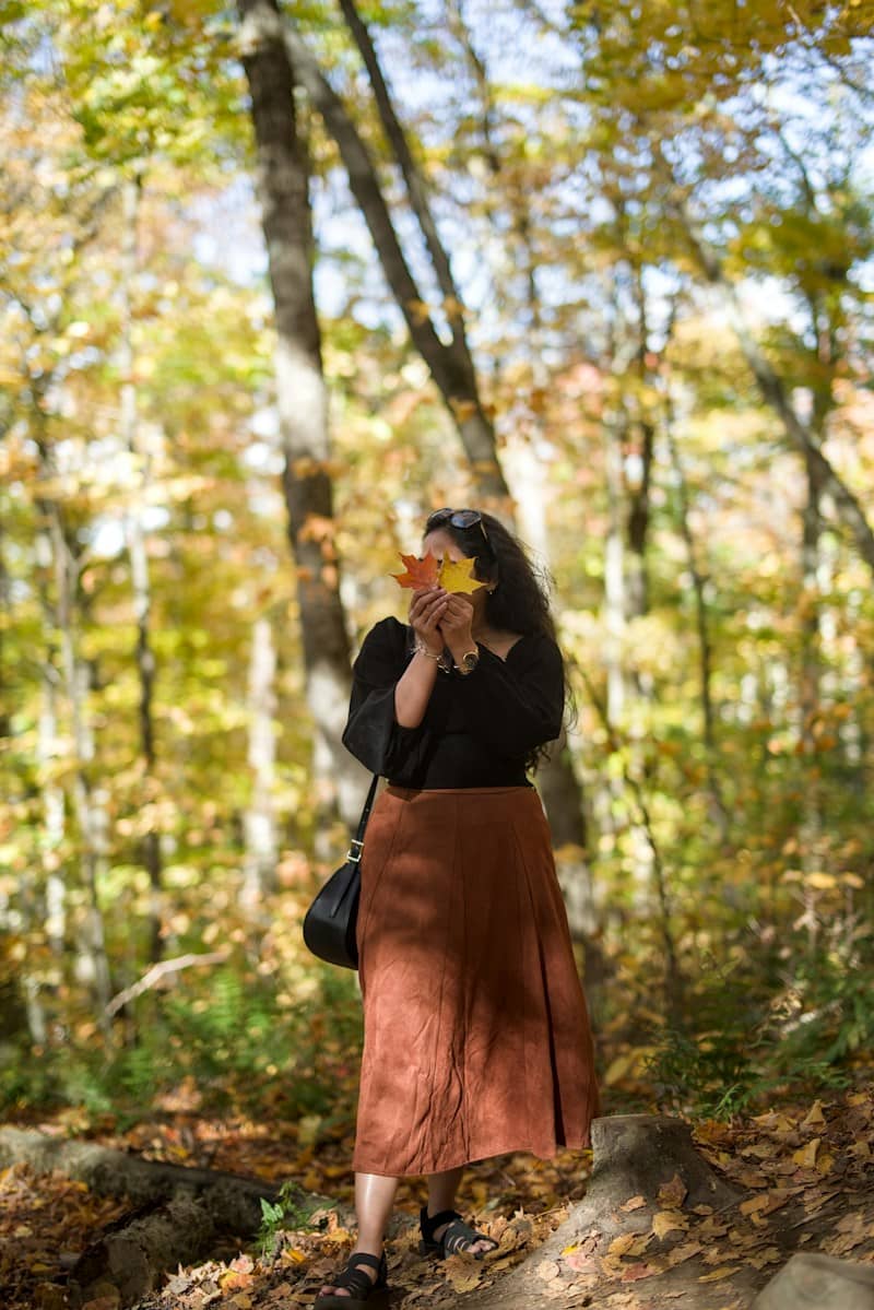 A woman in a black top and brown skirt holding autumn leaves over her face