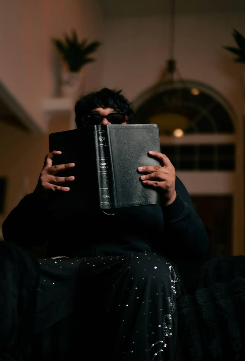 A person in dark clothing holding a black book in front of their face