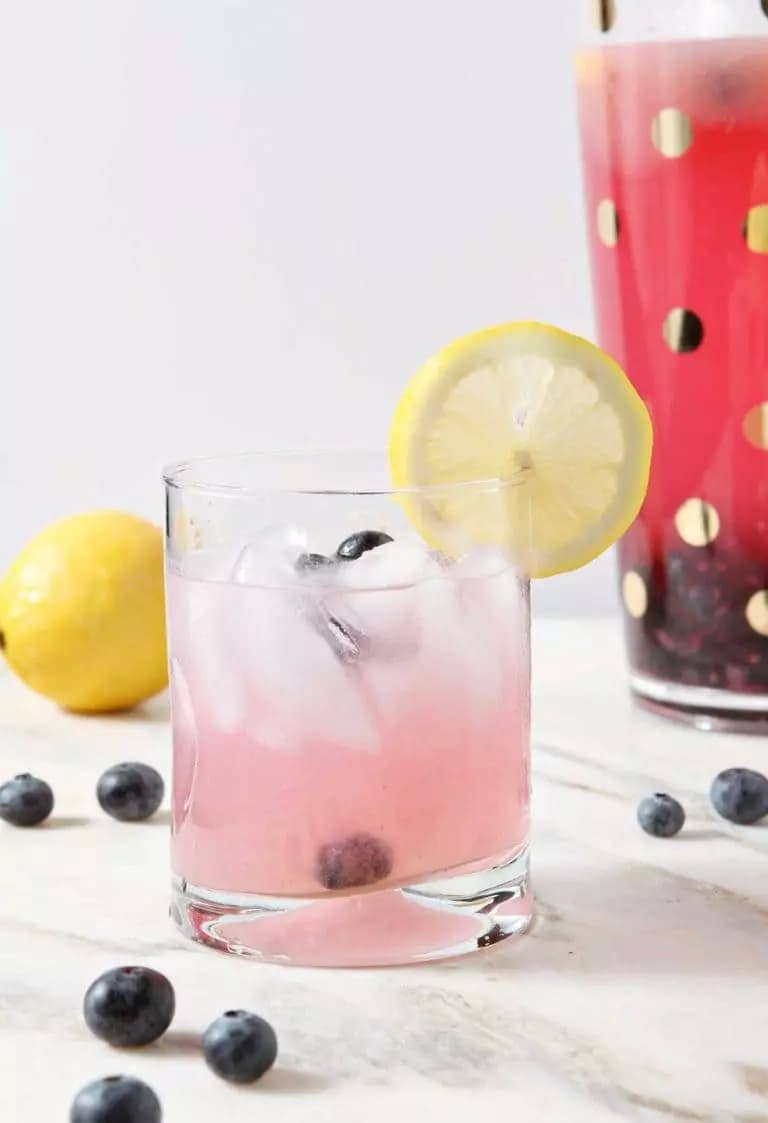 a glass of blueberry lemonade garnished with a lemon wheel on a marble surface