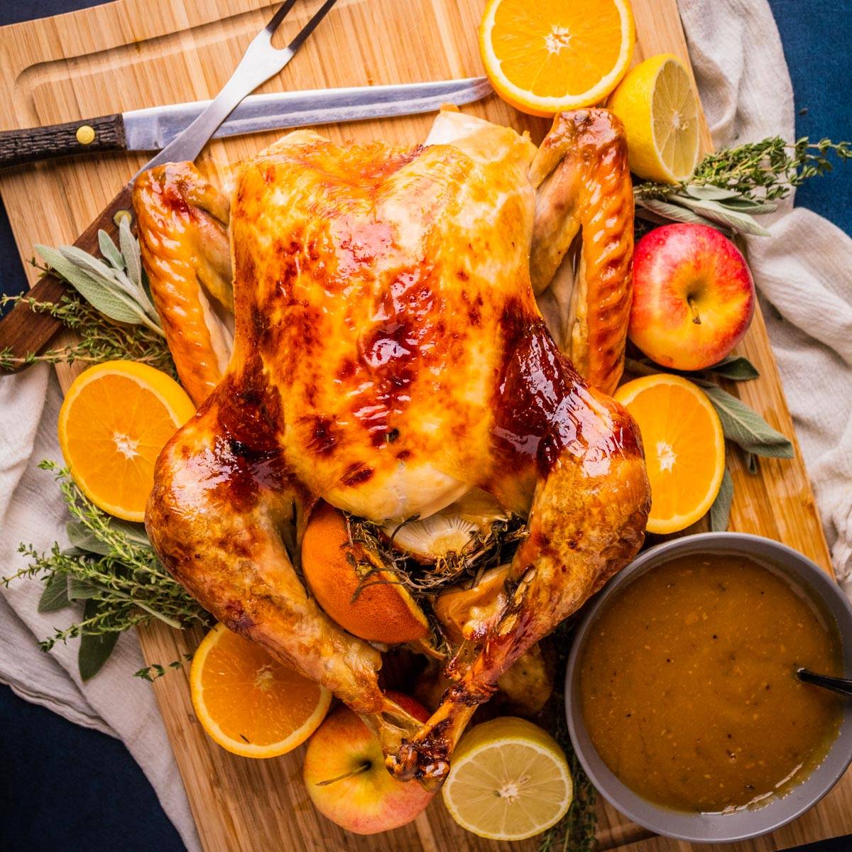 roasted turkey on a wooden cutting board with fresh herbs, fruit and gravy.