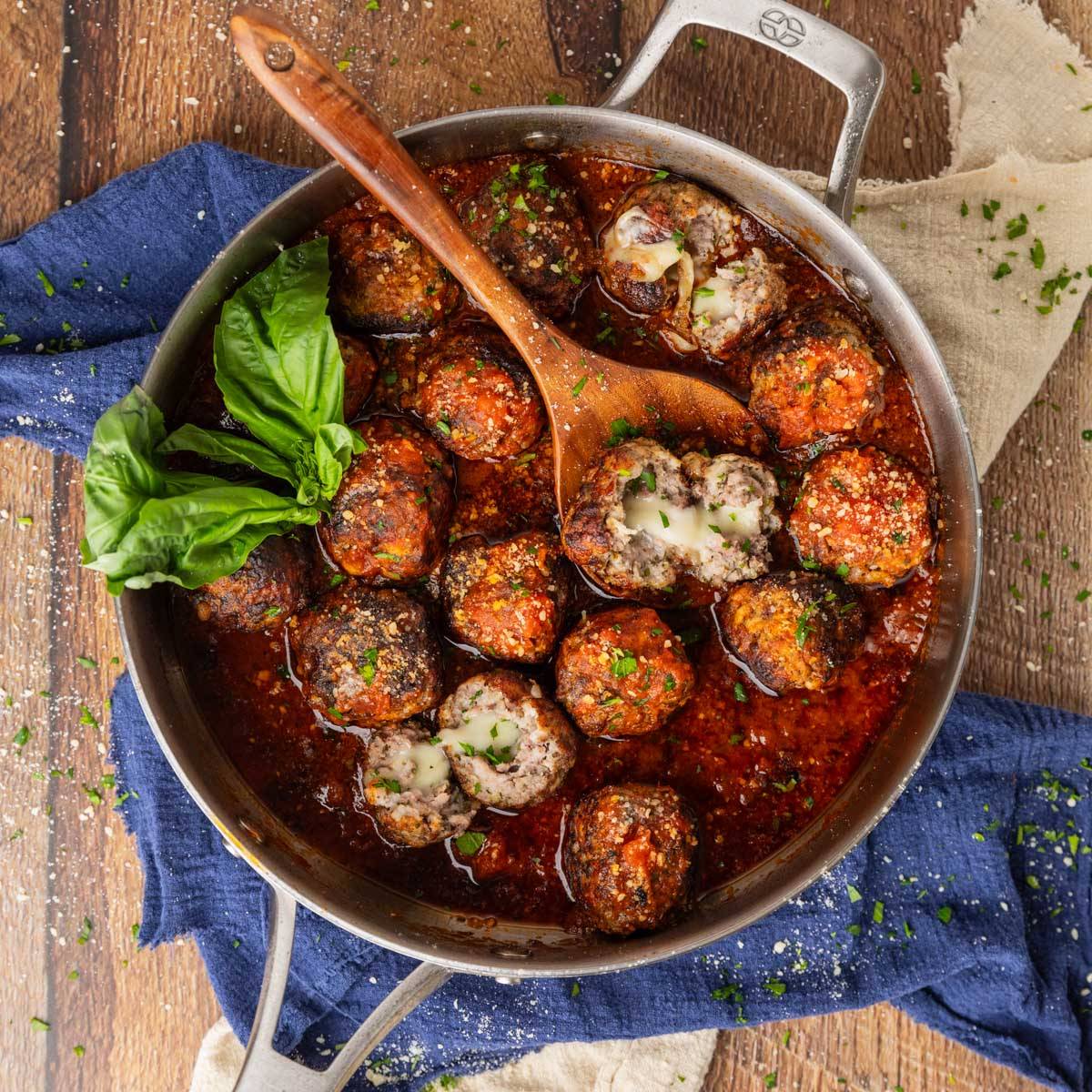 meatballs in red sauce in a skillet with a wooden spoon with some broken open to see melted mozzarella inside.